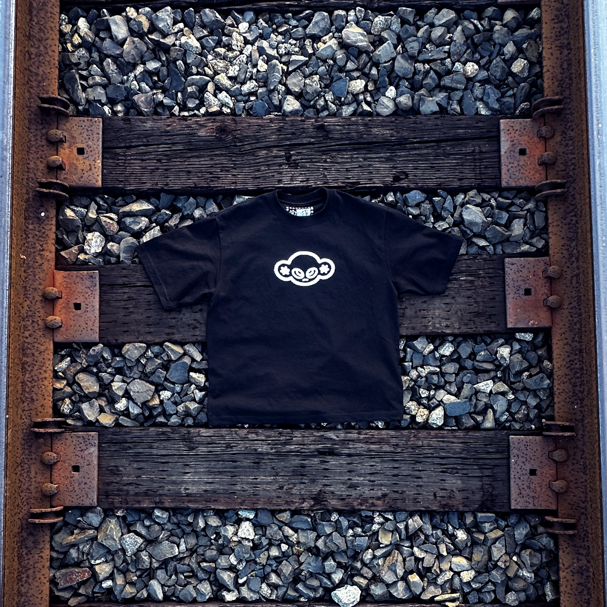 Black t-shirt with a white logo on a wooden train traack platform with gravel underneath