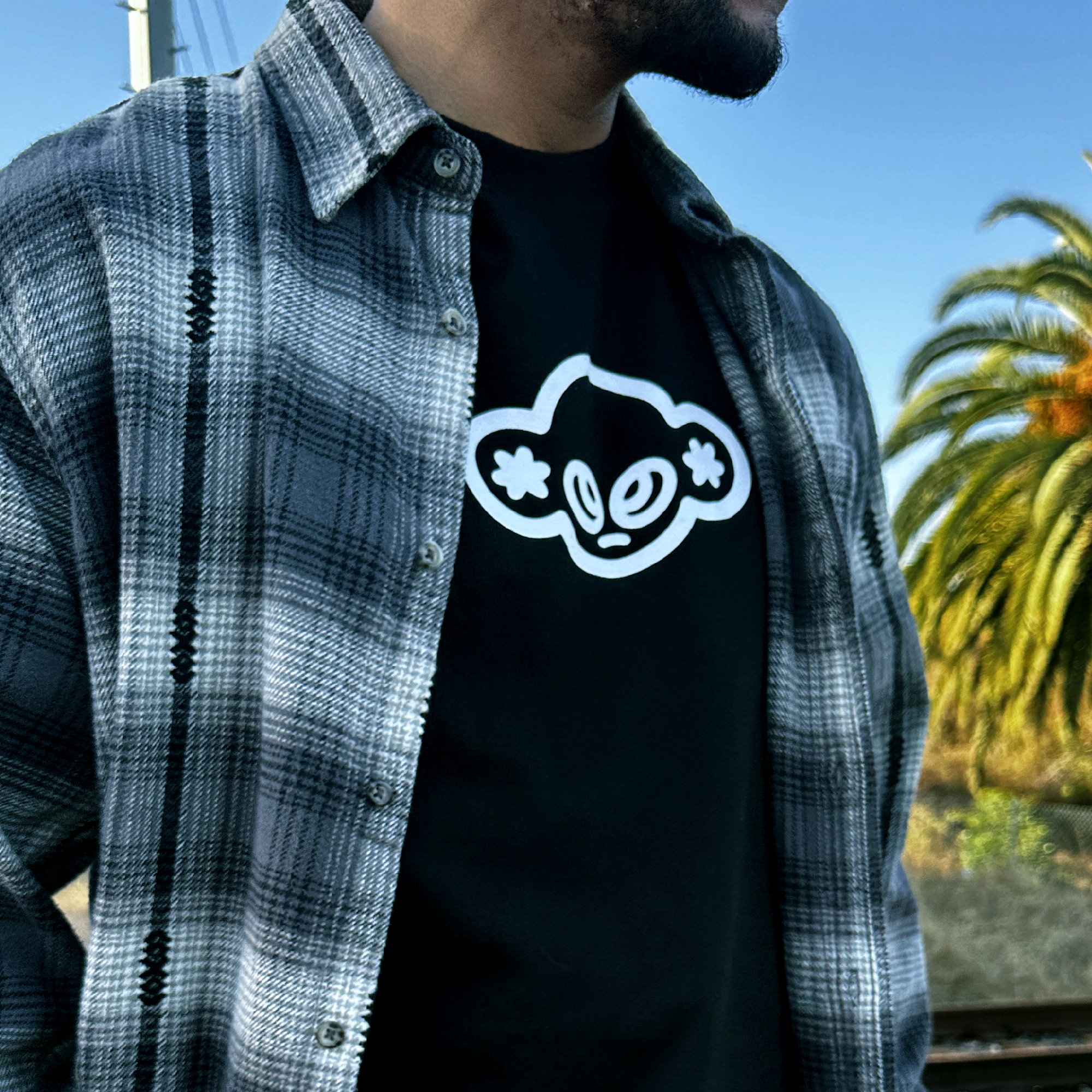 Person wearing a black t-shirt with a white logo and a plaid shirt outdoors.